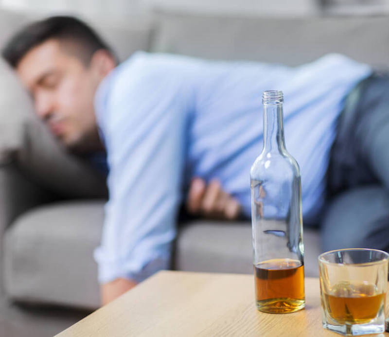 homme endormi dans une position anormale sur son divan avec une bouteille et un verre d'alcool en premier plan